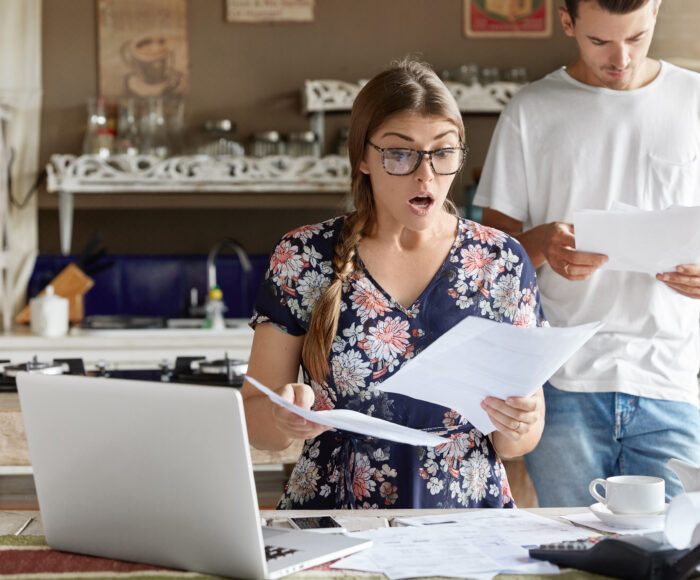 Young female looks with shocked expression into papers, recieve high taxes and had to pay much money for bills, sits against kitchen interior, her husband stands behinds, studies documents attentively